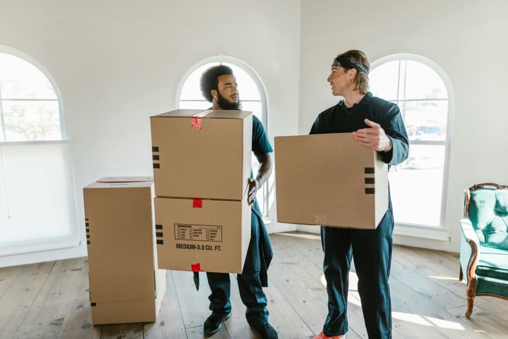 pexels-photo-7464244-7464244 Two professional movers carrying cardboard boxes inside a bright room with large windows.
